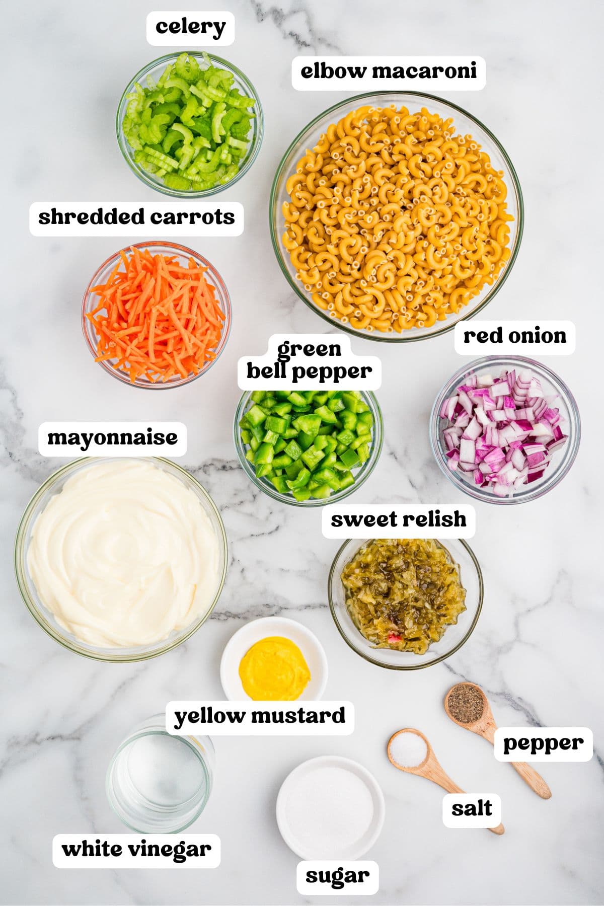 Top-down view of individual bowls containing labeled ingredients for macaroni salad: celery, elbow macaroni, shredded carrots, green bell pepper, red onion, mayonnaise, sweet relish, yellow mustard, white vinegar, sugar, salt, and pepper.