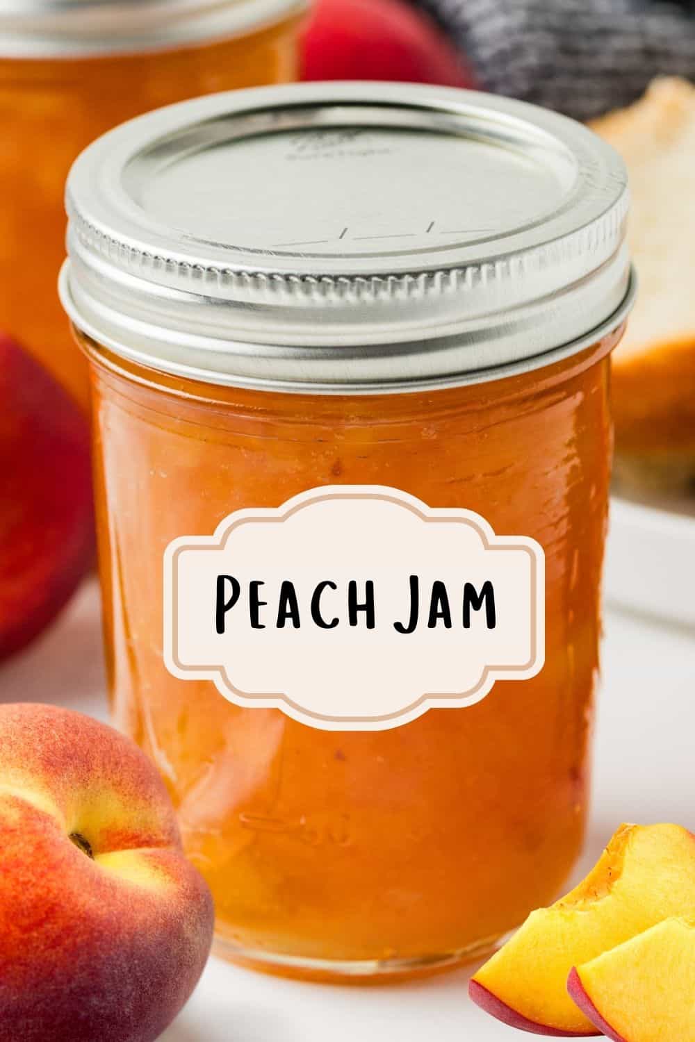 A sealed mason jar labeled “Peach Jam” sits on a marble surface surrounded by fresh peaches, a slice of bread, and another jar in the background.