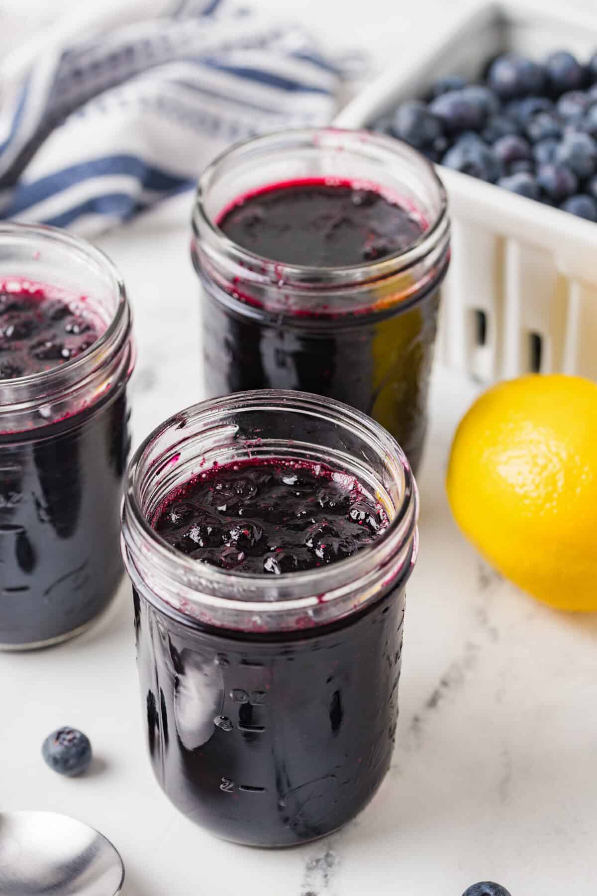 Three jars filled with homemade blueberry jam without pectin beside a lemon and fresh blueberries.