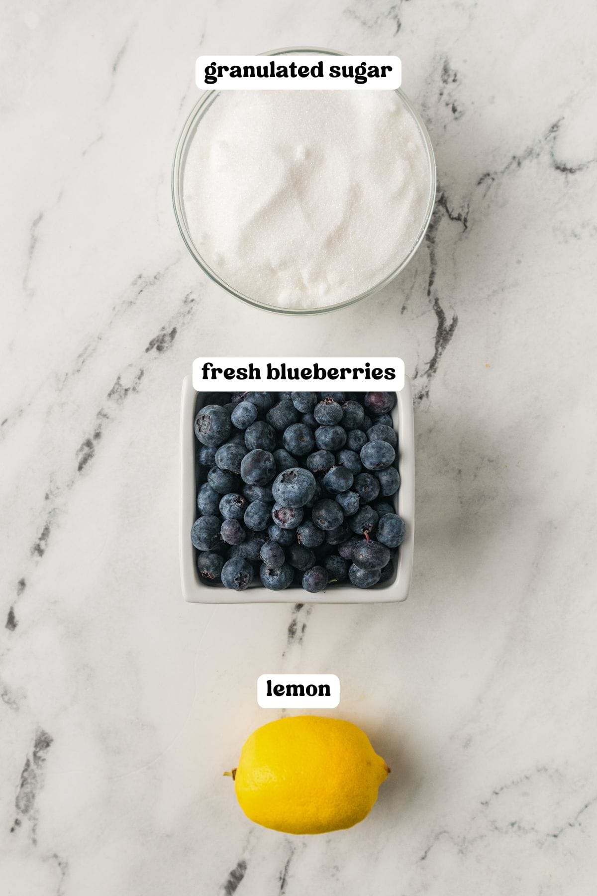 Overhead view of granulated sugar, fresh blueberries, and a lemon on a marble surface, each labeled.