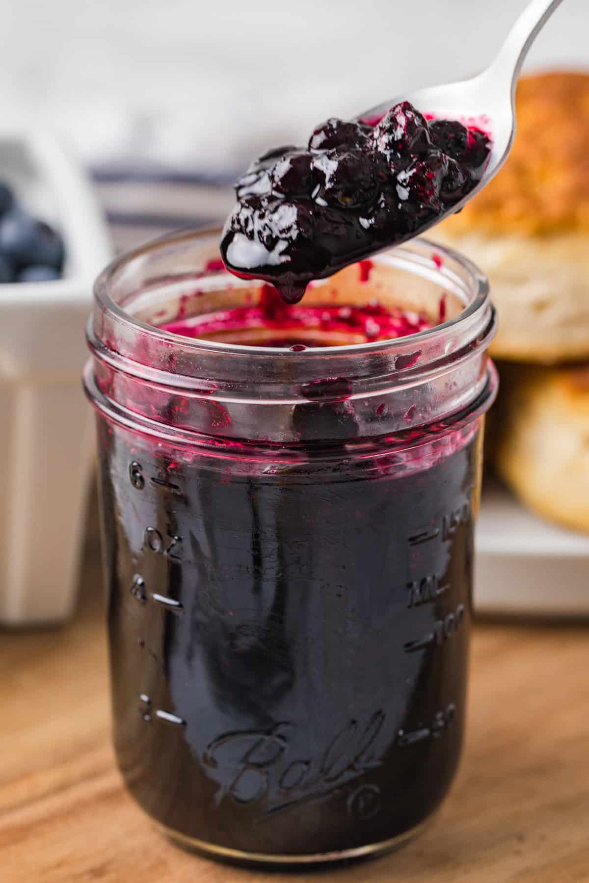 A close-up of a spoonful of thick blueberry jam being lifted from a mason jar. The jar sits on a wooden surface with biscuits and fresh blueberries in the background.