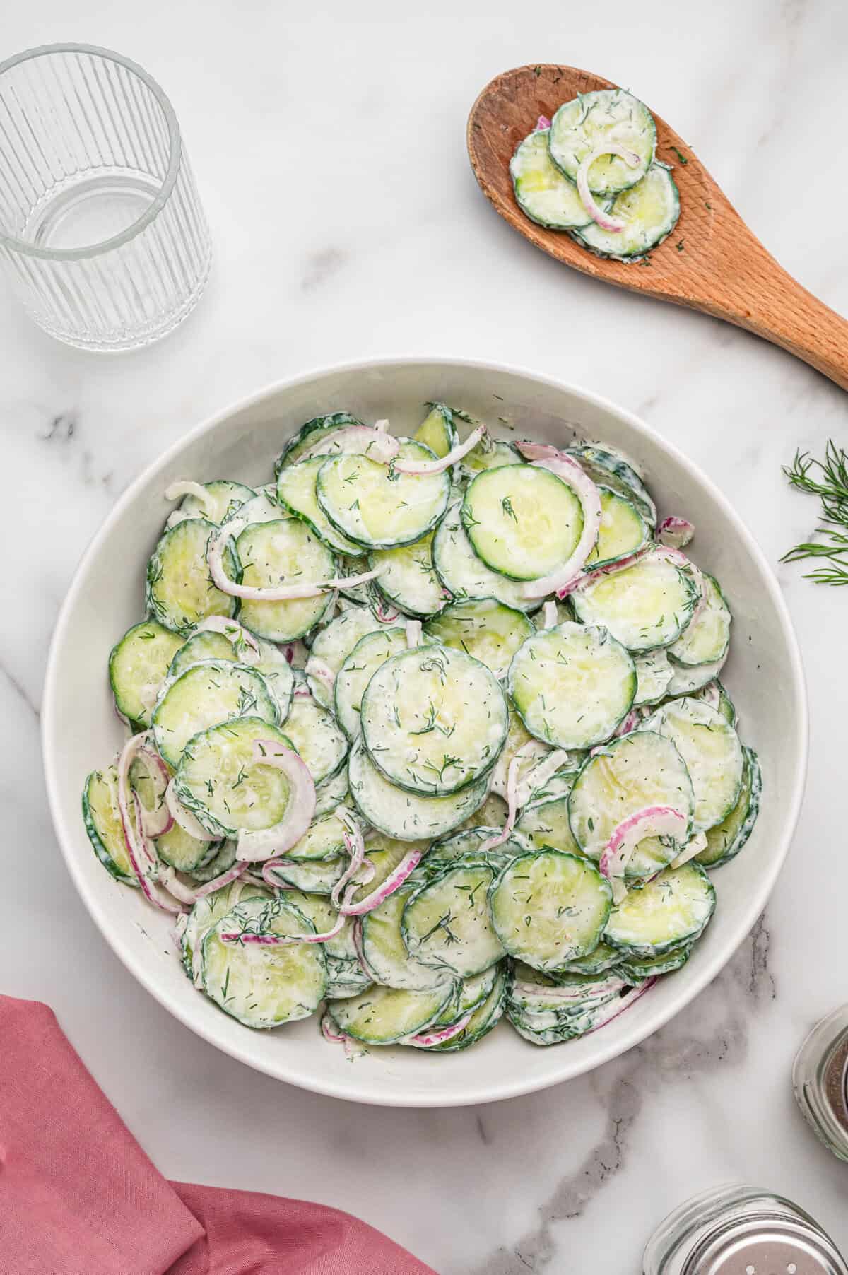 Bowl of creamy cucumber dill salad on a marble surface, with a wooden spoon holding more salad resting beside it.