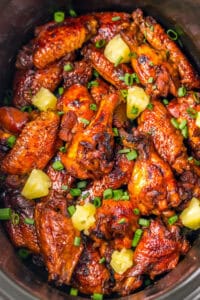 A crockpot packed with glazed Hawaiian wings. The chicken wings are topped with chunks of pineapple and chopped scallions.