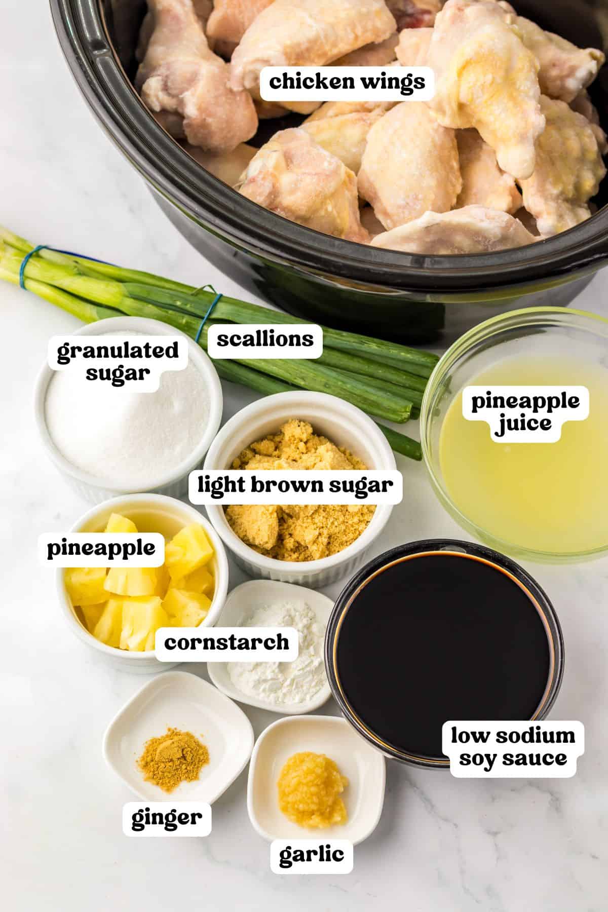Overhead view of labeled ingredients for Hawaiian chicken wings: raw wings in a crockpot, scallions, granulated sugar, light brown sugar, pineapple chunks, cornstarch, pineapple juice, soy sauce, garlic, and ground ginger.