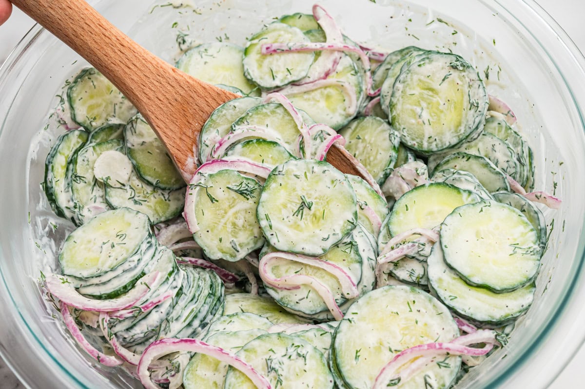 Wooden spoon stirring creamy cucumber salad in a glass bowl, showing cucumber slices, red onion, and creamy dill dressing.