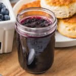 An open jar filled with homemade blueberry jam with biscuits and fresh blueberries in the background.