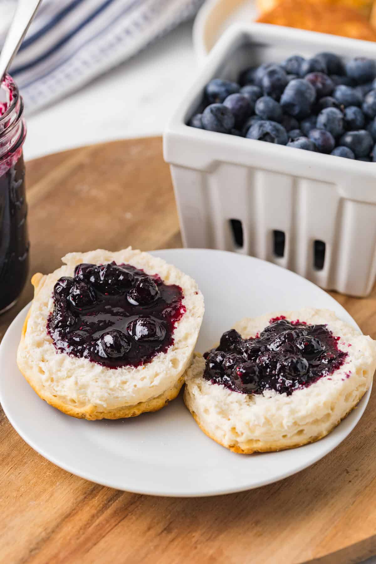 Biscuit halves on a white plate topped with blueberry jam. Fresh blueberries and homemade jar in mason jar are in background.