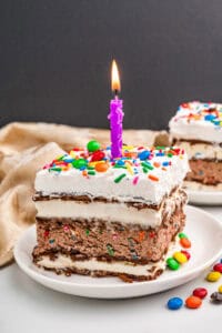 Ice cream sandwich cake slice topped with cool whip, sprinkles, mini M&Ms, and a lit purple candle.