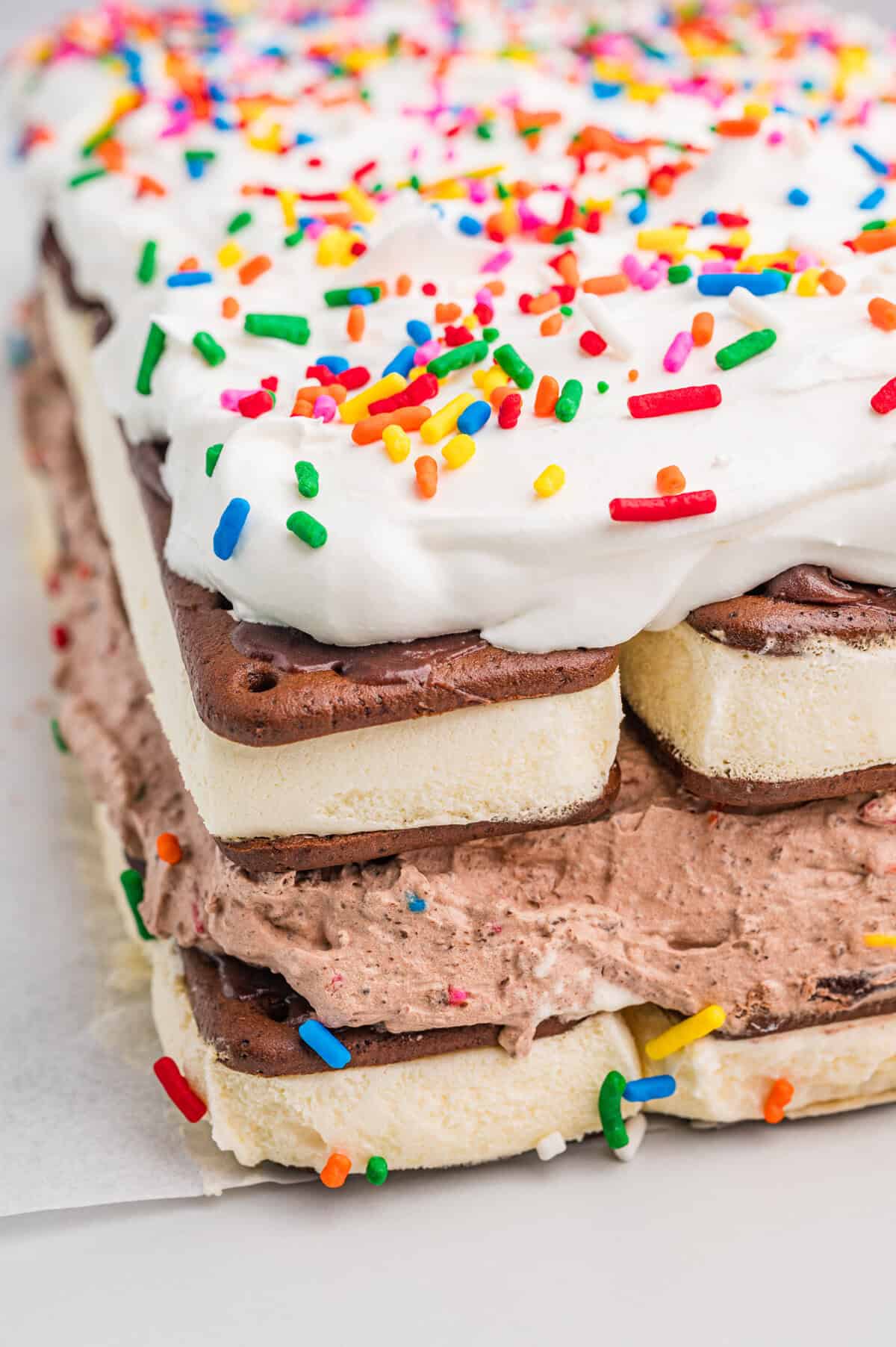 lose-up corner view of a layered ice cream sandwich cake. The cake consists of alternating layers of vanilla ice cream sandwiches and chocolate whipped filling with colorful sprinkles, topped with a thick layer of whipped topping and more rainbow sprinkles.