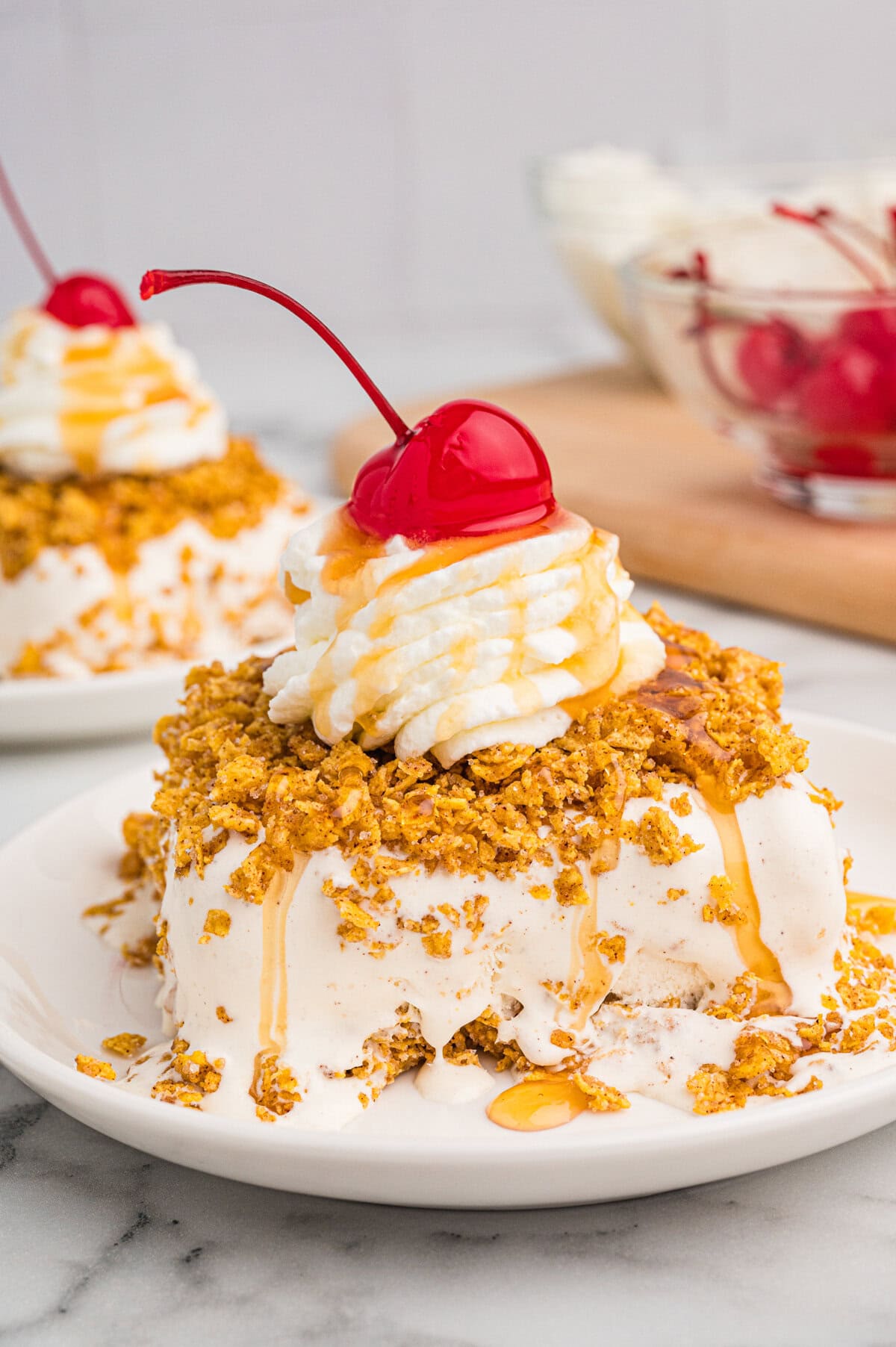 Fried ice cream dessert on a white plate, coated in a crispy golden crust, topped with whipped cream, caramel drizzle, and a maraschino cherry.
