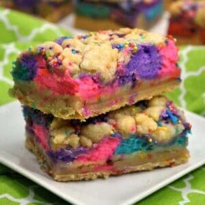 Two colorful layered dessert bars with bright pink, purple, and blue cheesecake swirls and cookie crumbles on top.