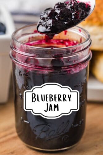 Close-up of a mason jar filled with blueberry jam, with a spoon lifting jam above it. Stylized white label on the jar reads “Blueberry jam.”