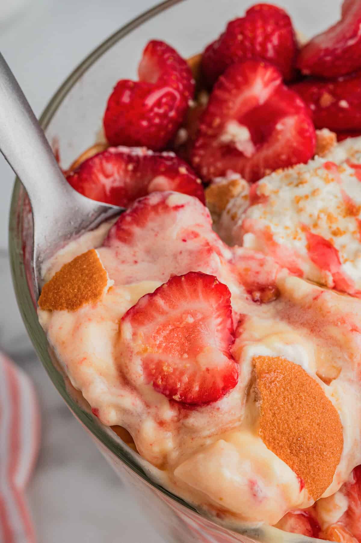 A spoon holding a portion of the strawberry banana pudding dessert with strawberries, whipped cream, pudding, and pieces of Nilla wafers, shown in a close-up view above the trifle bowl.