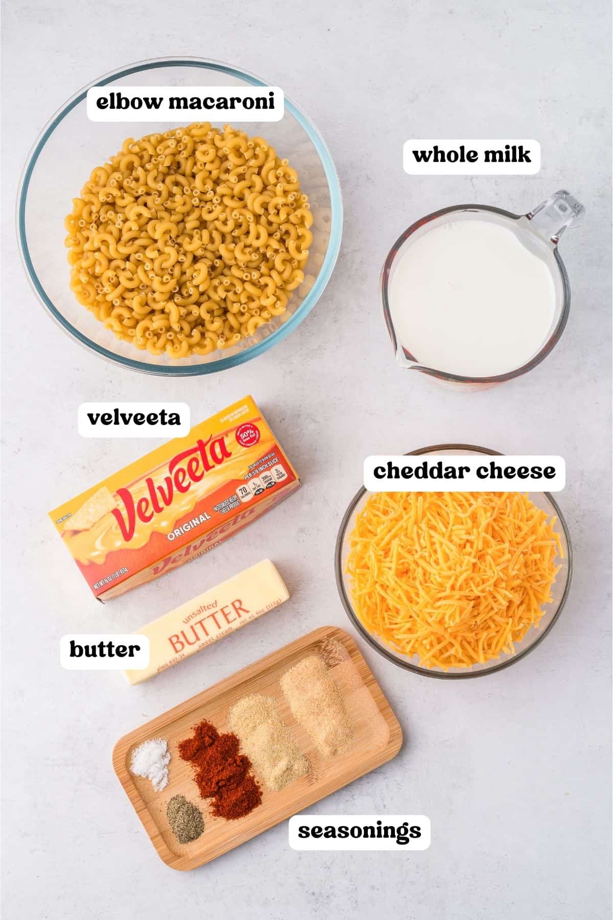 Overhead image showing labeled ingredients for slow cooker mac and cheese: elbow macaroni in a glass bowl, whole milk in a measuring cup, shredded cheddar cheese, a block of Velveeta, a stick of butter, and a small wooden tray with assorted seasonings.