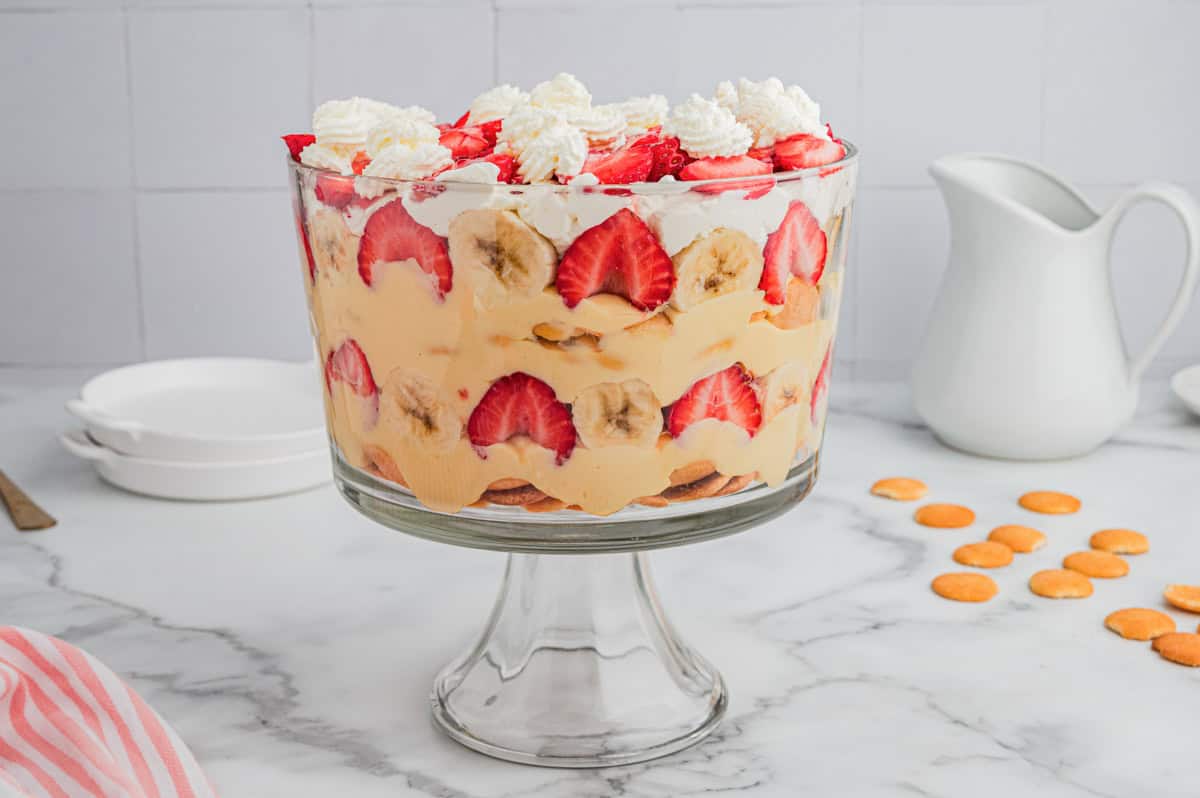 A glass trifle bowl filled with strawberry banana pudding, showing layered rows of sliced strawberries, bananas, vanilla pudding, whipped cream, and Nilla wafers, with whipped cream dollops and strawberries on top.