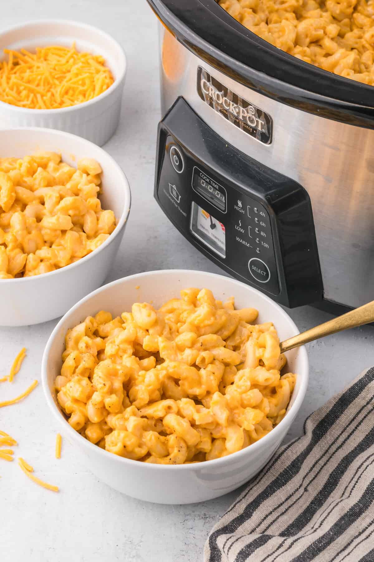 Two bowls of creamy homemade macaroni and cheese next to a stainless steel Crock Pot filled with more slow cooker mac and cheese.