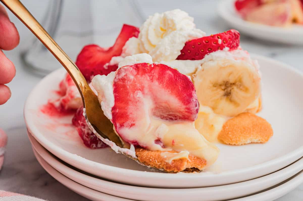 A close-up of a spoon lifting a bite of strawberry banana pudding from a white plate, showing layers of sliced strawberries, banana, vanilla pudding, whipped cream, and a Nilla wafer on the bottom.