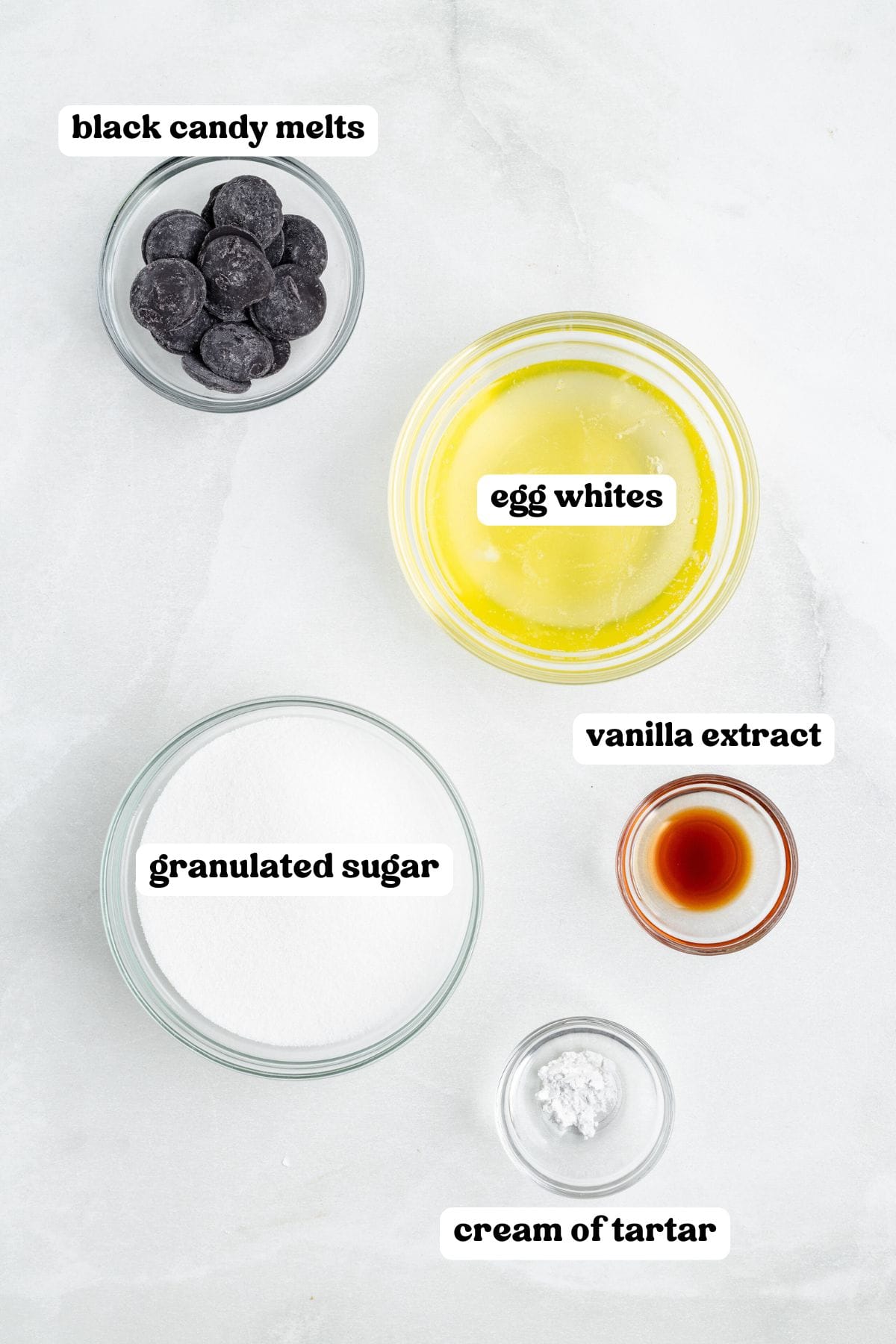 Overhead view of labeled ingredients in clear bowls including: black candy melts, egg whites, granulated sugar, vanilla extract, and cream of tartar.