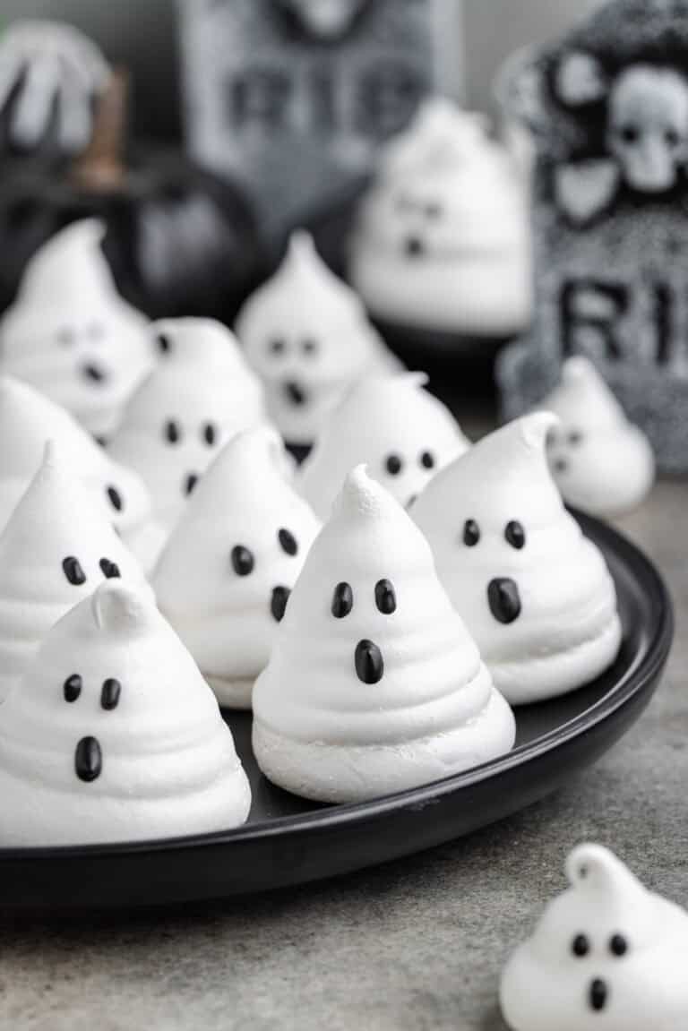 Ghost meringues with black candy eyes and mouths are arranged on a plate. The background includes Halloween-themed tombstone decorations and black pumpkins, creating a festive setting.