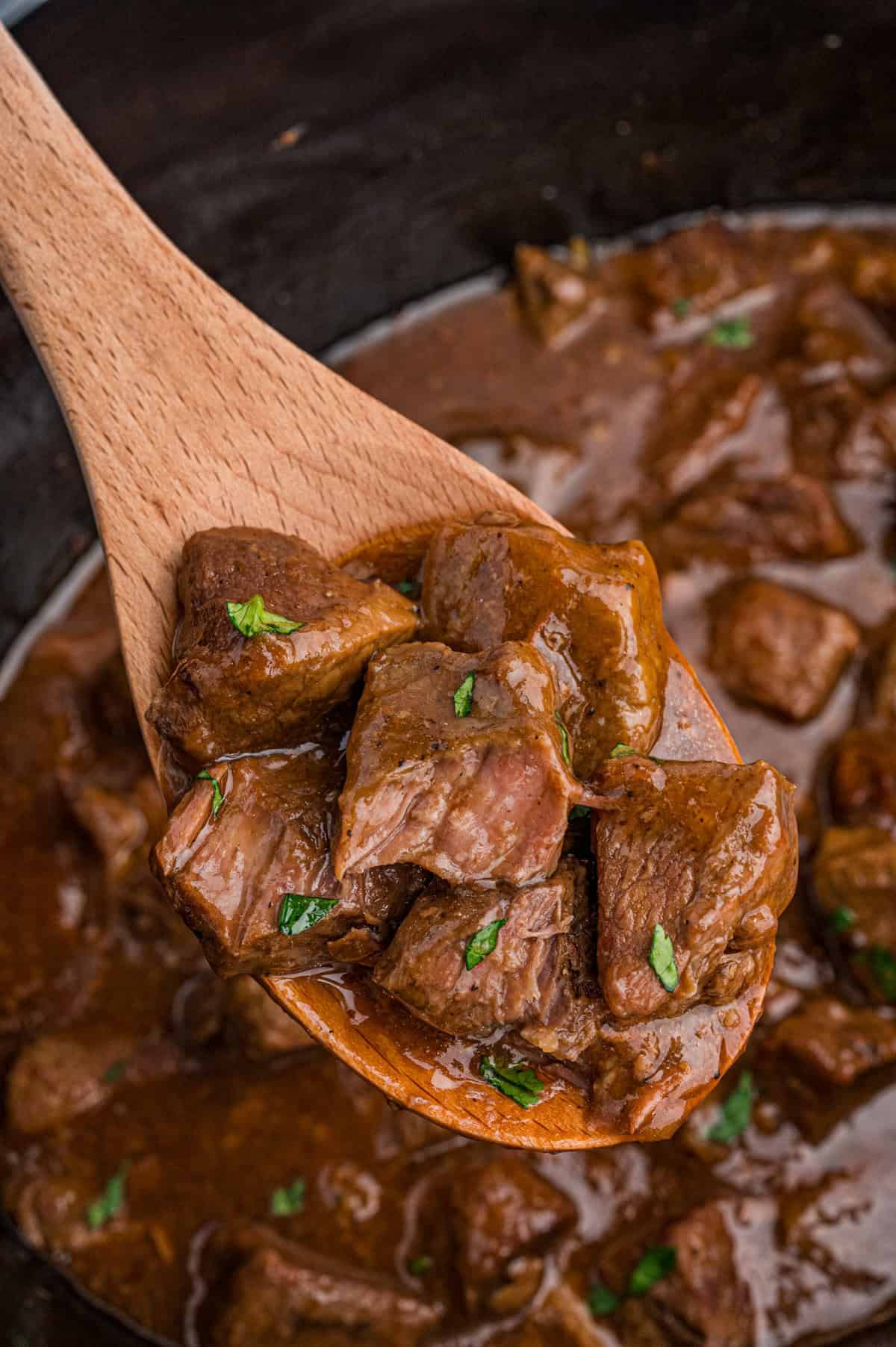 A wooden spoon lifting several glossy, honey bourbon steak tips garnished with parsley from a crockpot filled with more steak bites.