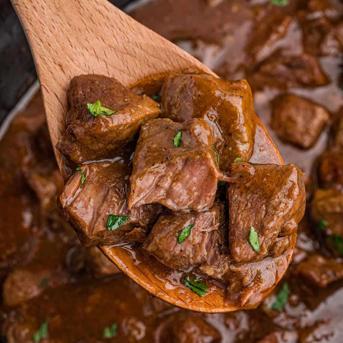 A wooden spoon lifting honey bourbon steak tips garnished with parsley from a slow cooker filled with more steak bites.