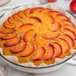 A peach upside-down cake in a parchment-lined white round dish. The top of the golden cake is decorated with arranged slices of fresh peach in a circular pattern.