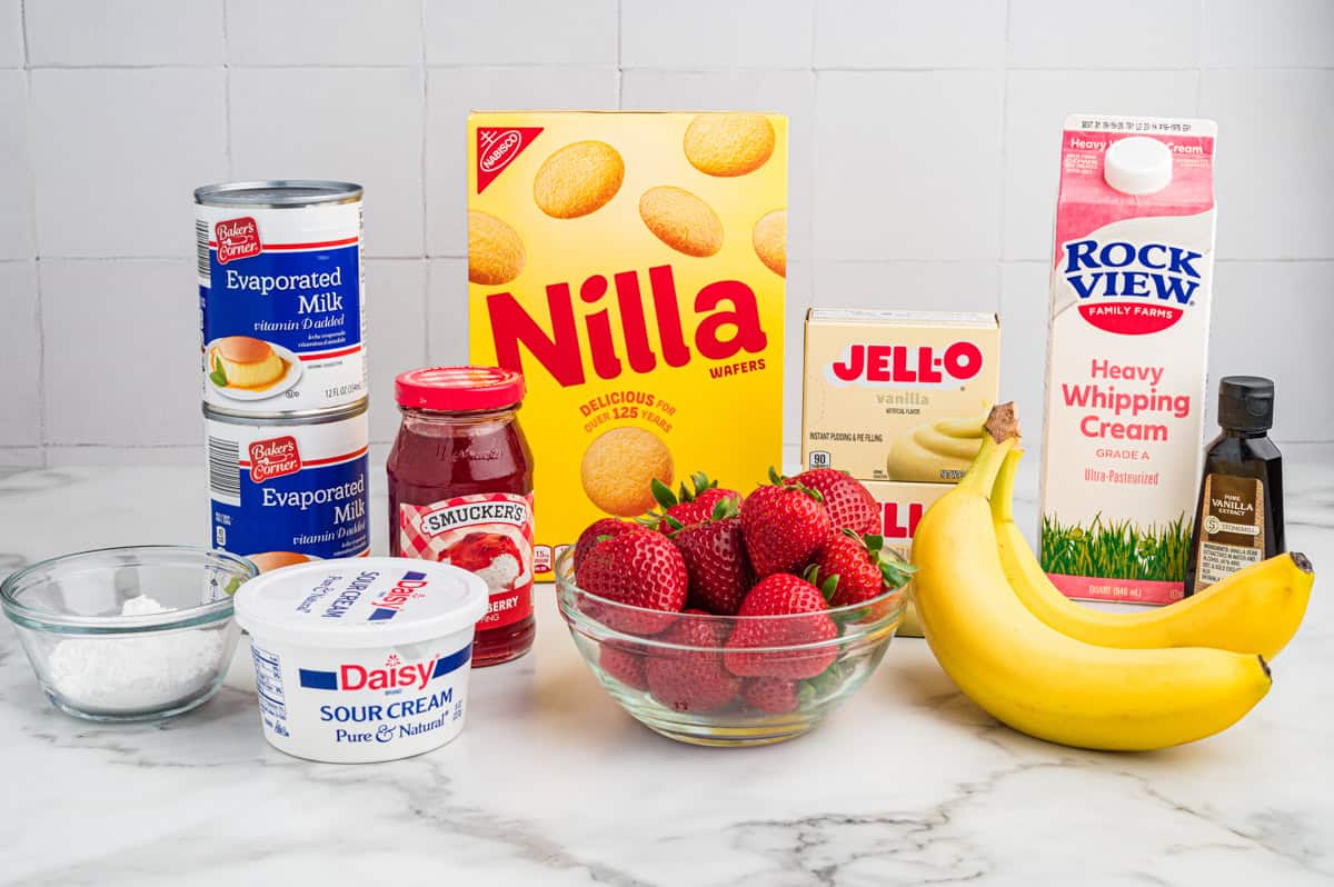 Ingredients for making strawberry banana pudding, including fresh strawberries, bananas, Nilla wafers, vanilla pudding mix, heavy whipping cream, sour cream, evaporated milk, vanilla extract, powdered sugar, and strawberry syrup.