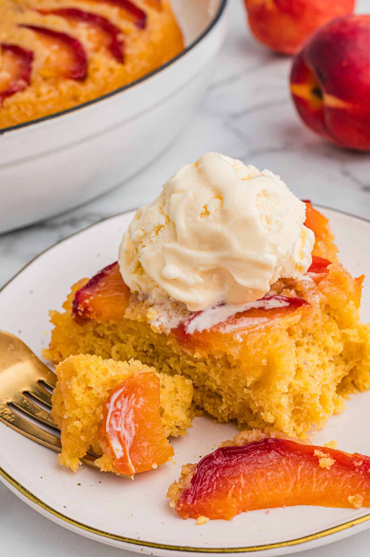 A slice of golden peach upside-down cake with visible peach slices on top, served on a plate and topped with a scoop of vanilla ice cream. A fork with a bite of cake rests nearby.