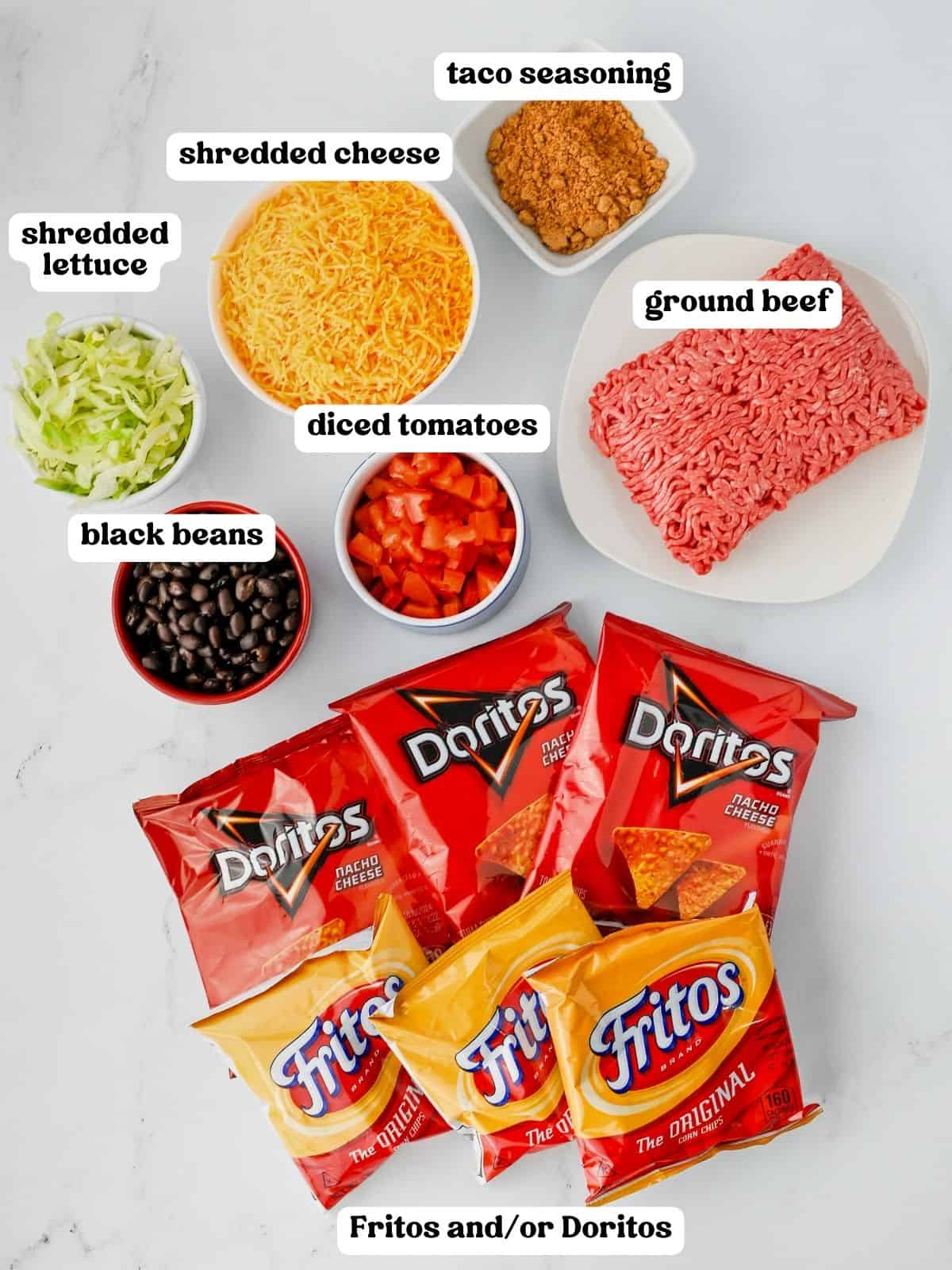 Walking taco ingredients including shredded cheese, taco seasoning, raw ground beef, shredded lettuce, black beans, diced tomatoes, and several unopened bags of Doritos and Fritos.