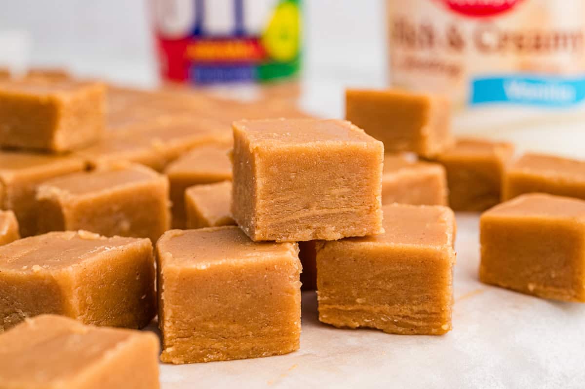Several cubes of peanut butter fudge resting on parchment paper. A Jif peanut butter jar and a can of frosting are blurred in the background.