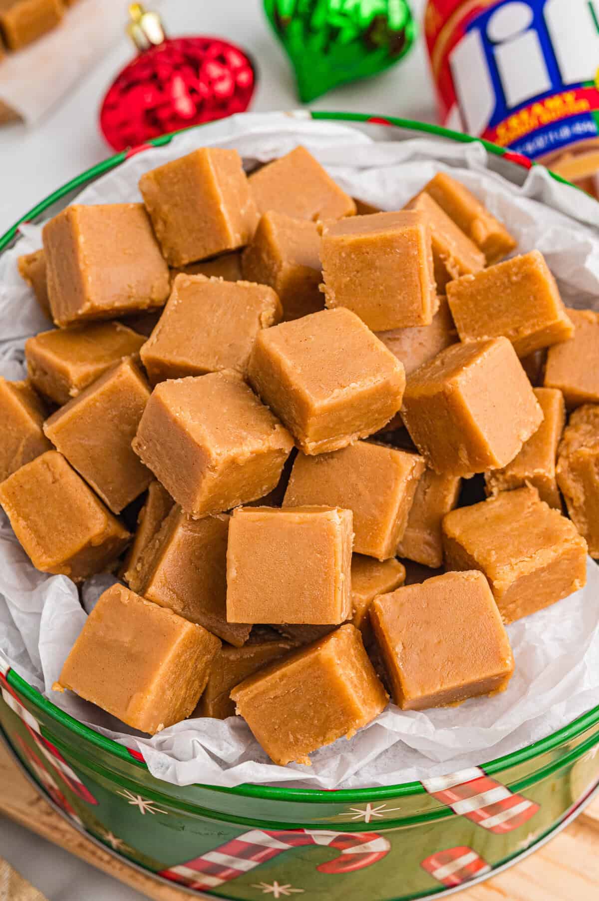 Peanut butter fudge pieces in a holiday tin. Christmas ornaments and a jar of Jif peanut butter are visible in the background.
