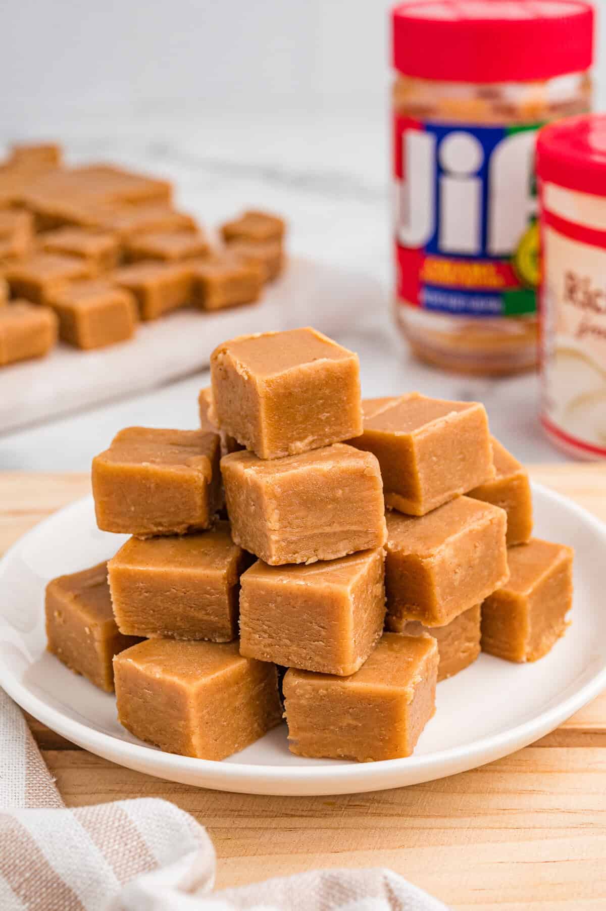 Stack of smooth, light brown peanut butter fudge cubes arranged on a white plate. In the background, jars of Jif peanut butter and Betty Crocker vanilla frosting are slightly blurred, with more fudge pieces visible on parchment paper.