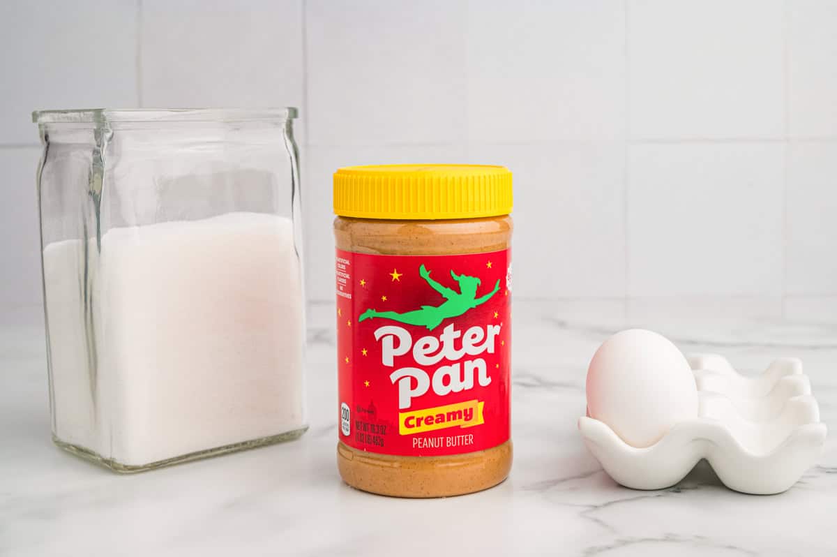 A jar of Peter Pan creamy peanut butter, a glass container of granulated sugar, and a single white egg in a ceramic egg holder.