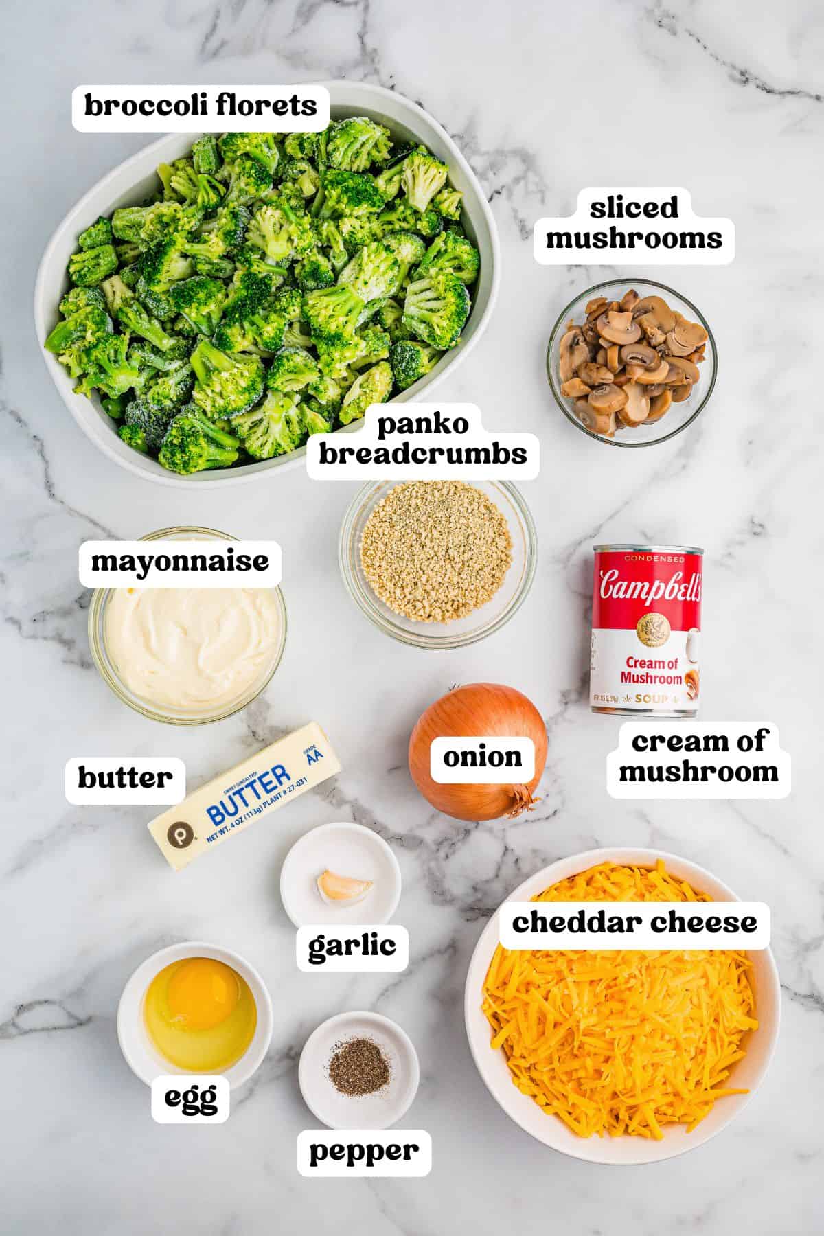 Overhead view of labeled ingredients for broccoli casserole including broccoli florets, sliced mushrooms, panko breadcrumbs, mayonnaise, butter, onion, cream of mushroom soup, garlic, egg, pepper, and shredded cheddar cheese.