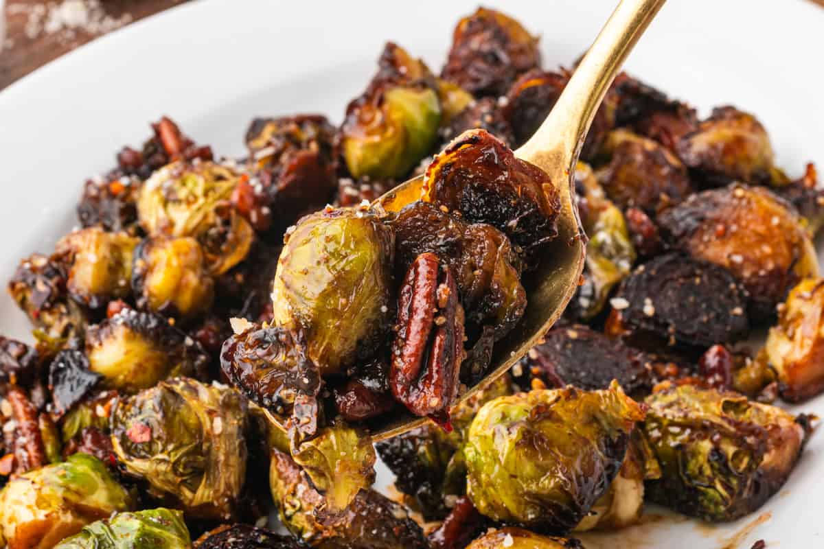 Golden spoon lifting a serving of caramelized Brussels sprouts and pecans, coated in a glossy glaze. The plate beneath is full of additional roasted sprouts and darkened bits from caramelization.