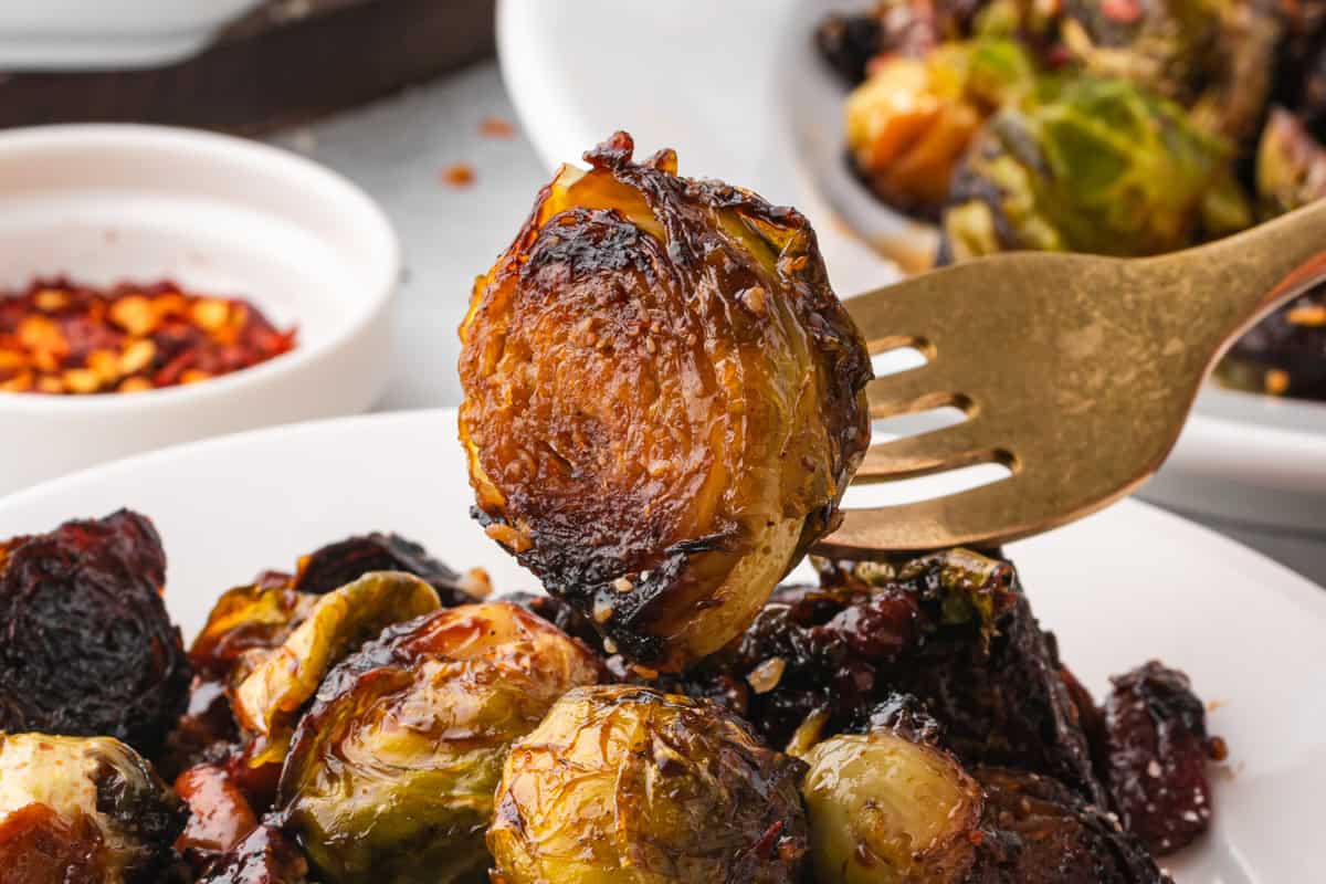 Close-up of a fork holding a single caramelized Brussels sprout with a crisp, browned surface. The background shows more Brussels sprouts on a plate and a small bowl of red pepper flakes.