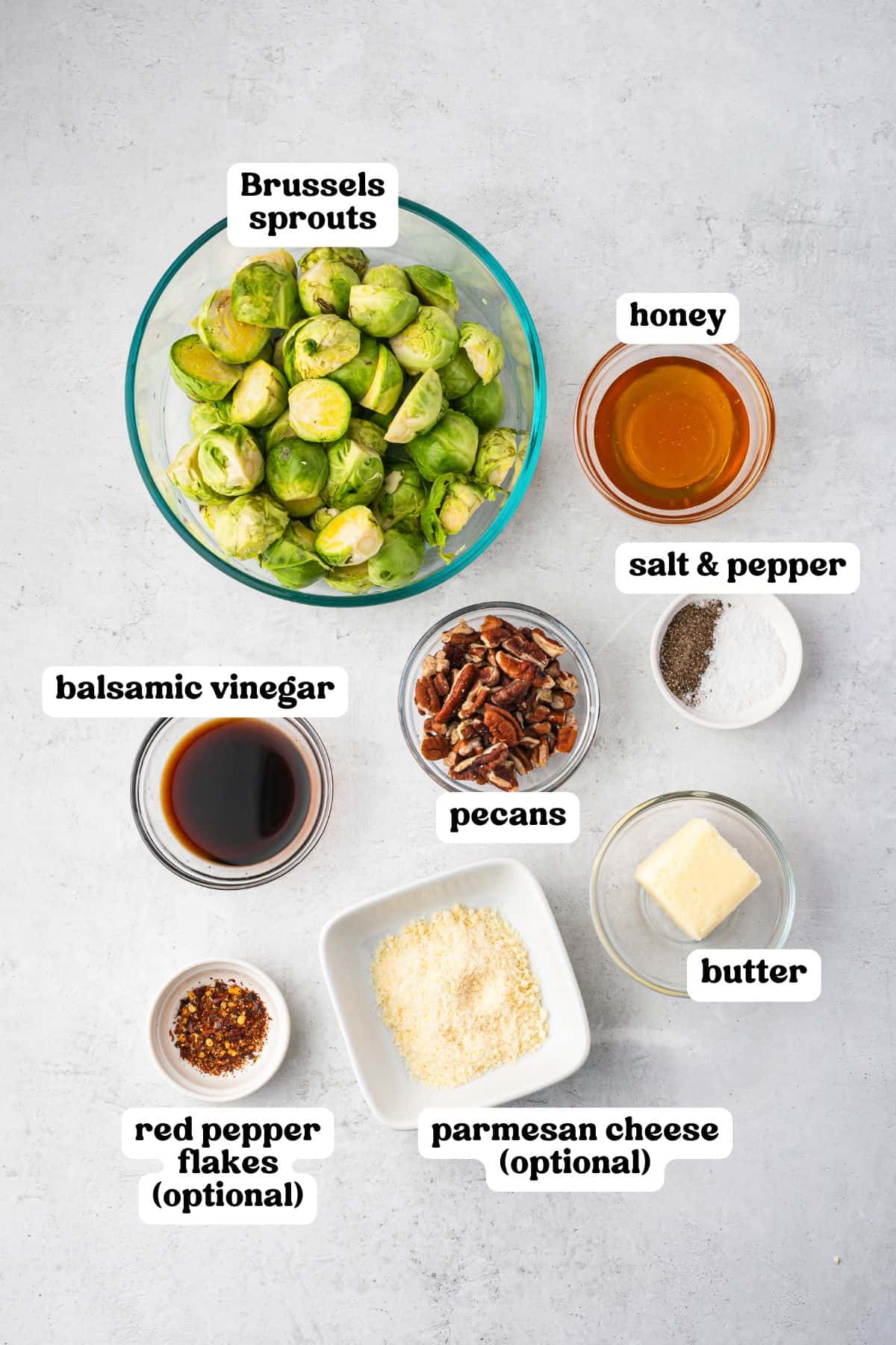 Overhead image of all ingredients labeled: halved Brussels sprouts in a glass bowl, honey, salt and pepper, balsamic vinegar, chopped pecans, butter, red pepper flakes (optional), and parmesan cheese (optional), all arranged on a light surface.