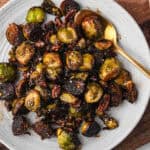 A plate filled with caramelized Brussels sprouts and pecans, sprinkled with seasoning. A gold spoon rests in the dish.