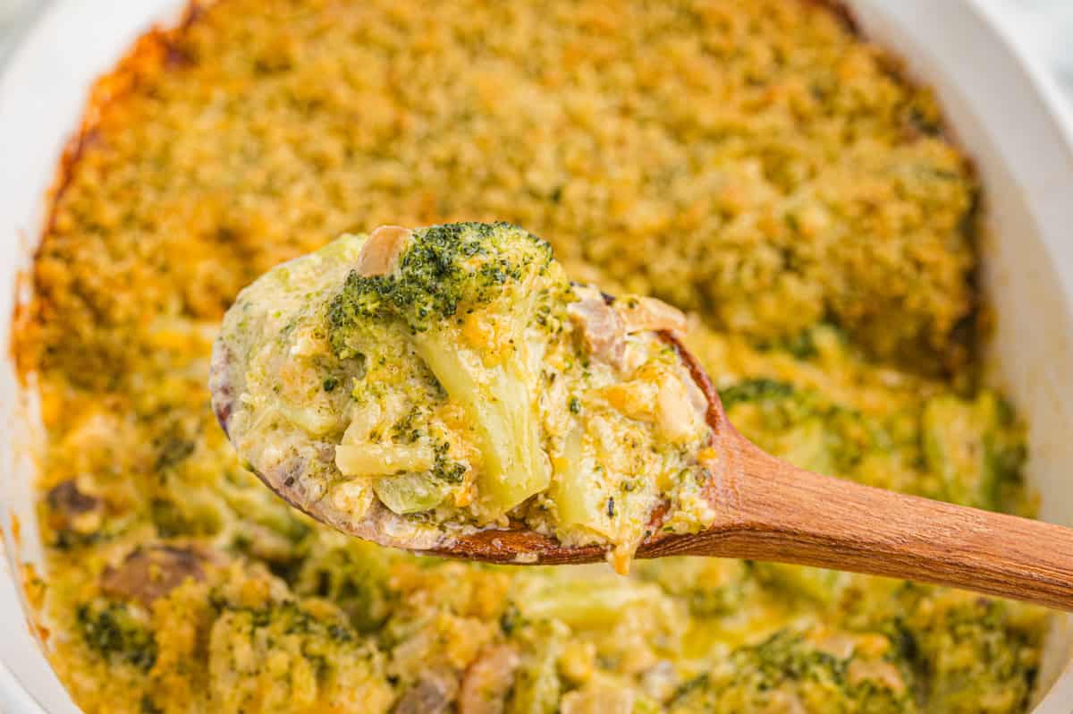 A wooden spoon lifts a creamy portion of broccoli casserole above the baking dish, showing broccoli, mushrooms, and melted cheese and onion beneath a crispy topping.