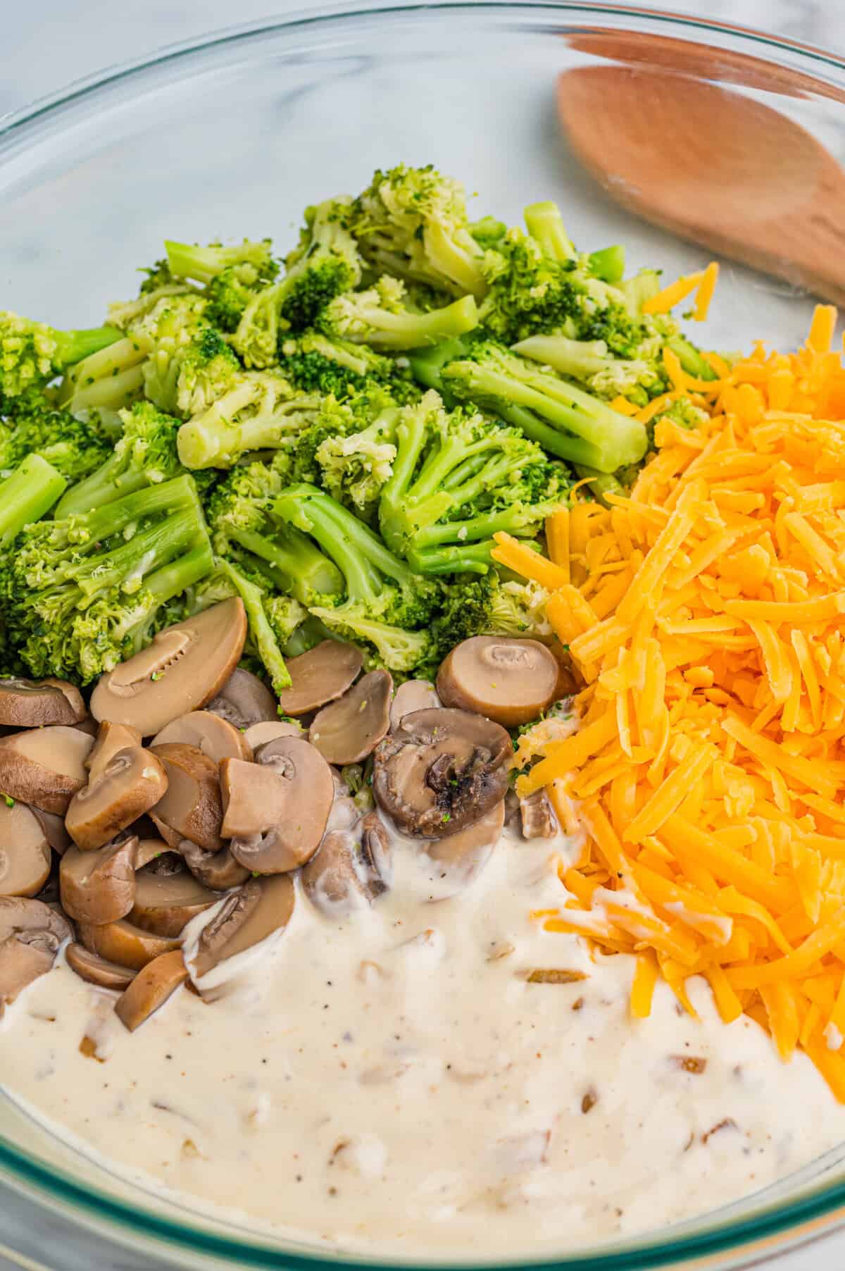 Mixing bowl containing bright green broccoli florets, sliced mushrooms, shredded cheddar cheese, and creamy mushroom soup, ready to be mixed for casserole.