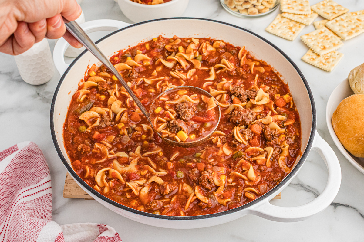 A ladle scooping hamburger soup from a large pot surrounded by crackers and rolls. The soup contains ground beef, egg noodles, corn, peas, green beans, carrots, and diced tomatoes.