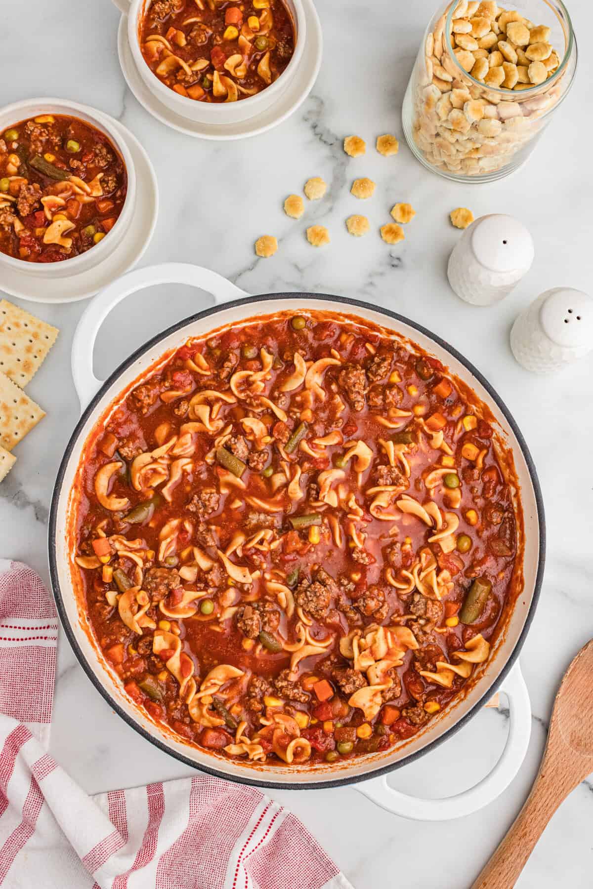 Top-down image of a white pot filled with hamburger noodle soup, featuring ground beef, egg noodles, mixed vegetables, and tomato-based broth. Two smaller bowls with soup are nearby, along with a glass jar of oyster crackers, scattered crackers, salt and pepper shakers, and saltine crackers.