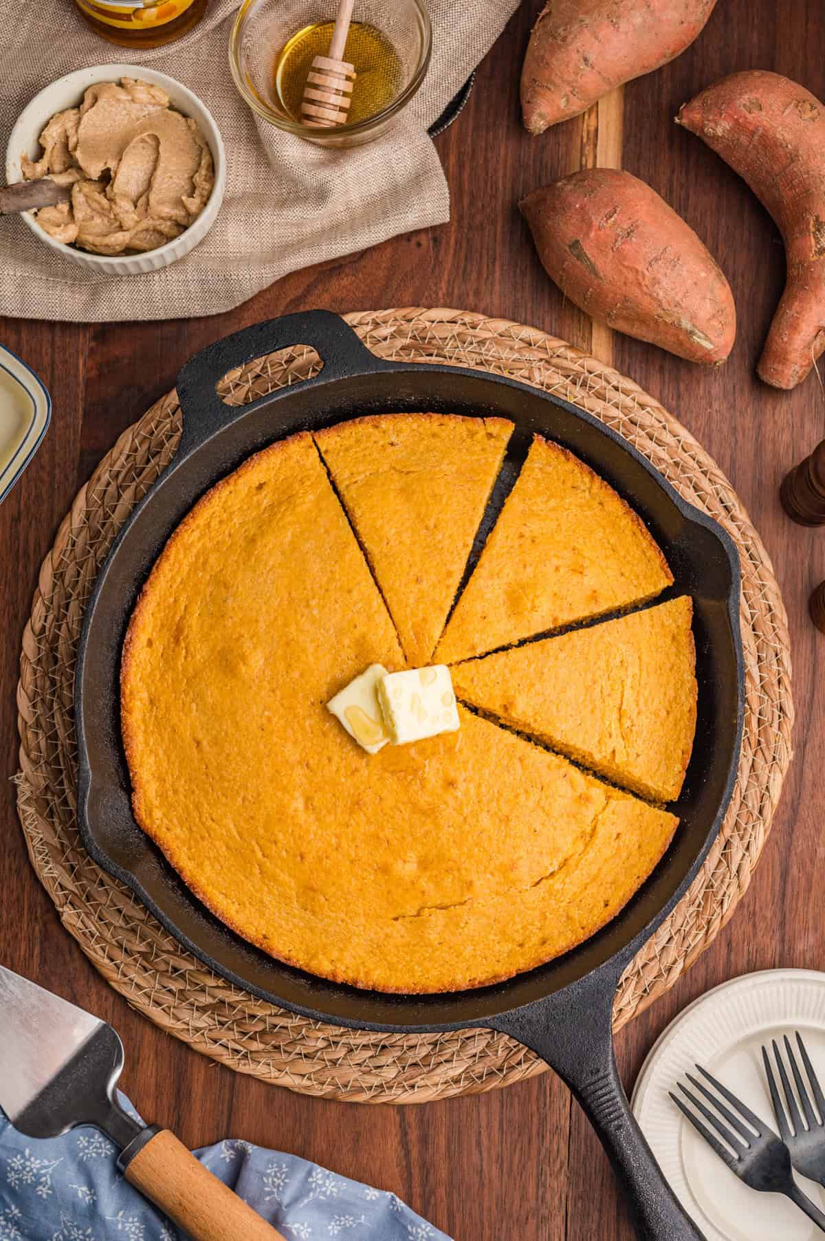 Overhead view of a cast iron skillet filled with sweet potato cornbread, sliced into wedges. A pat of butter is melting in the center, surrounded by sweet potatoes, a honey jar, cinnamon butter, and a serving spatula on a wooden table