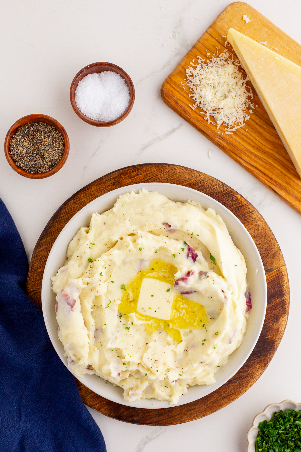 Overhead photo of a white bowl of mashed potatoes with red skin potatoes, garnished with a pat of melting butter and herbs, set on a wooden board with bowls of salt and pepper, a wedge of Parmesan cheese, and shredded cheese on a wooden cutting board.