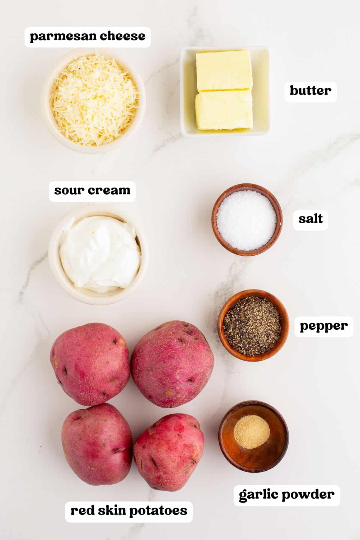 Overhead view of ingredients for mashed red potatoes with labels: Parmesan cheese, butter, sour cream, salt, pepper, garlic powder, and four whole red skin potatoes.