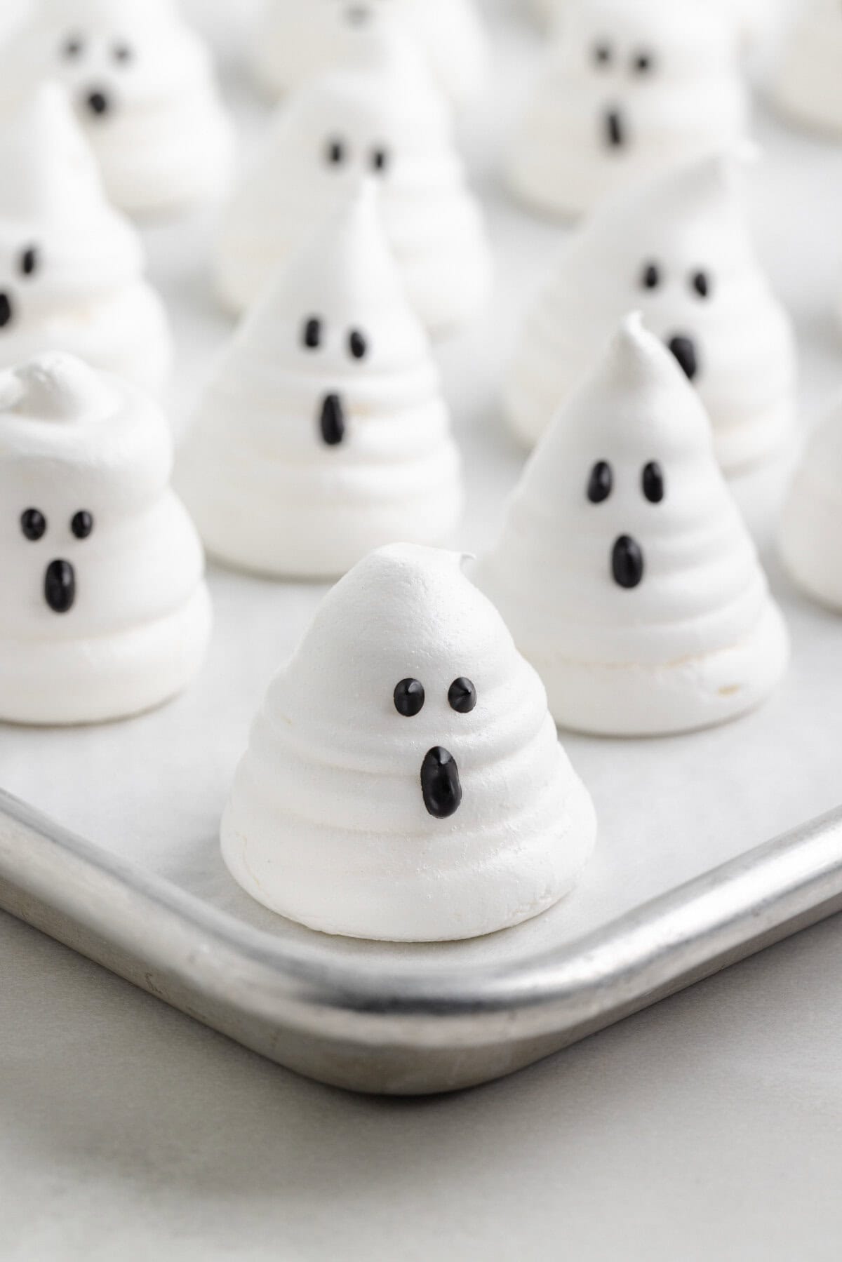 Baked meringue ghosts with black candy melt eyes and oval-shaped mouths arranged on a parchment-lined tray.