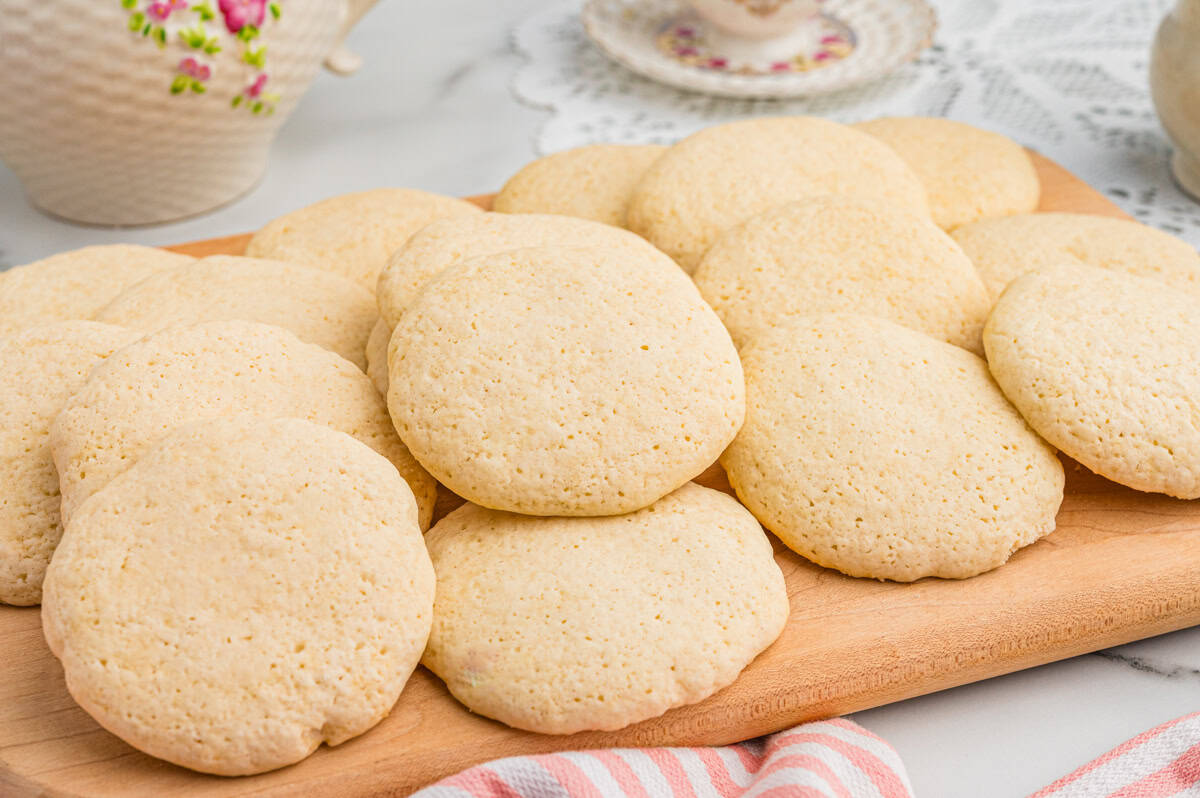 Angled view of old-fashioned tea cakes stacked and layered on a wooden board. A floral teapot and teacup sit in the background on a lace doily, suggesting a tea-time setting.