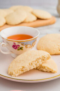 Close-up of a broken old-fashioned tea cake on a ceramic plate, revealing its soft interior. A floral-patterned teacup filled with tea sits behind it, with more cookies in the background.