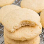Stacked old fashioned tea cakes with a visible bite out of top cookie revealing a soft, crumbly interior. The cookie sits on a cooling rack with more cookies in the background.