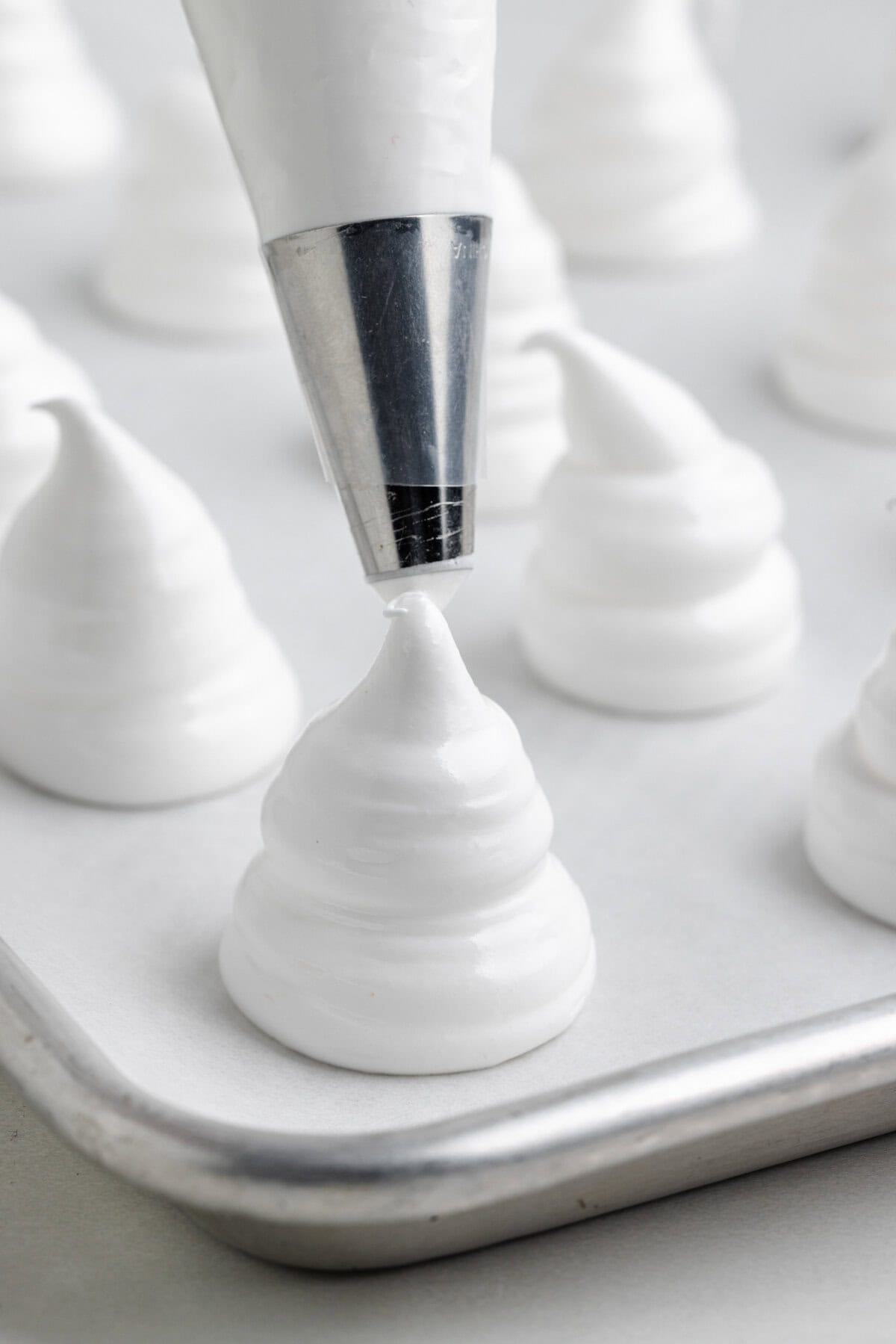 A piping tip forms a peaked swirl of white meringue on a parchment-lined baking tray.